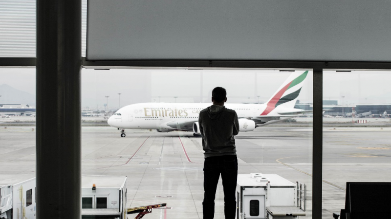 A man standing alone in an airport looking at a plane about to take off